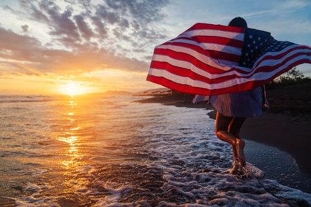 Happy woman holding american USA flag and running on the beach. Independence Day, travel conceptの写真素材