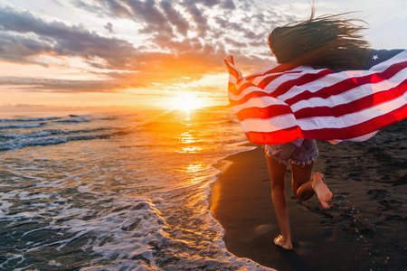 Happy woman holding american USA flag and running on the beach. Independence Day, travel conceptの写真素材