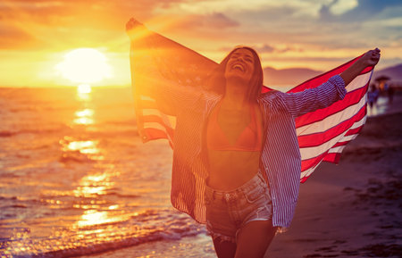 Happy woman holding american USA flag and running on the beach. Independence Day, travel conceptの写真素材