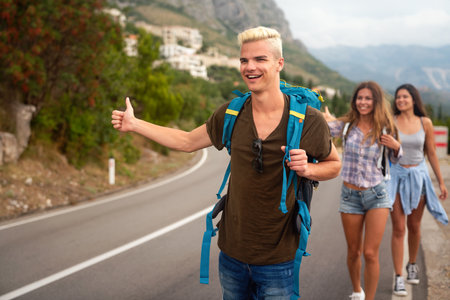 Cheerful interracial friends standing on road showing hitchhiking gestures. Tourism travelingの写真素材