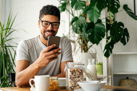 Young black man eating breakfast and reading the news online. Handsome man using mobile phoneの写真素材
