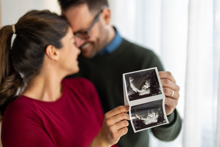 Happy young couple, pregnant woman, holding and showing ultrasound scan picture at homeの写真素材