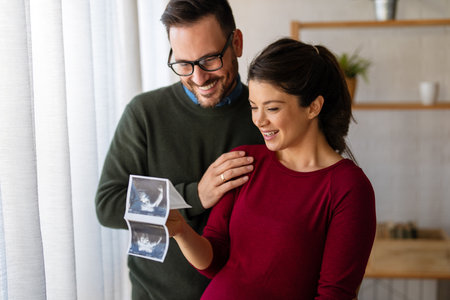 Happy young couple, pregnant woman, holding and showing ultrasound scan picture at homeの写真素材