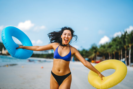 Young woman enjoys the warmth of the sun holding inflatable rings, celebrating the joy of beach lifeの写真素材