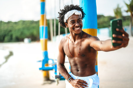 Smiling young black man taking a selfie on a vibrant beach, embracing the freedom of a digital nomadの写真素材