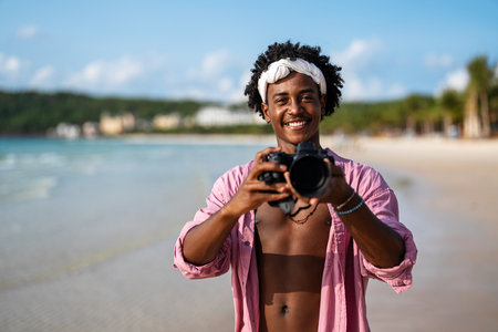 Joyful black man embraces the freedom of beach life, enjoying the fresh ocean breeze with his cameraの写真素材