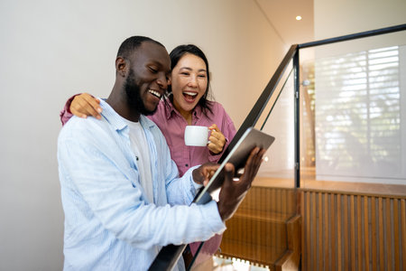 Multinational couple enjoying their morning routine, smiling and using laptop and tablet at homeの写真素材
