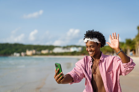 Happy young black man talking on his smartphone at a tropical beach, enjoying vacation, remote workの写真素材