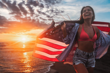 Beautiful patriotic young woman with the American flag held in her standing in the summer sunsetの写真素材