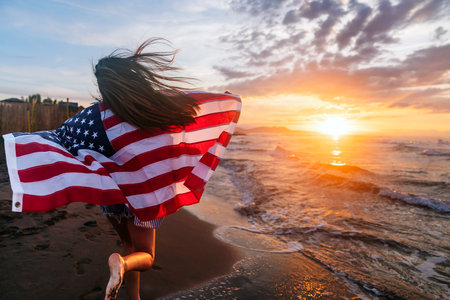 Beautiful patriotic young woman with the American flag held in her standing in the summer sunsetの写真素材
