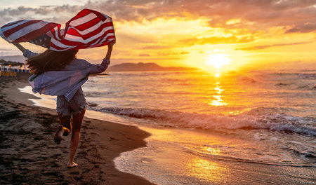 Beautiful patriotic young woman with the American flag held in her standing in the summer sunsetの写真素材