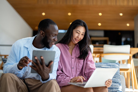 Multinational couple enjoying their morning routine, smiling and using laptop and tablet at homeの写真素材