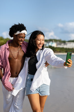 Diverse couple connected, embracing technology and social media while enjoying moments at the beachの写真素材