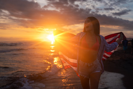 Beautiful patriotic young woman with the American flag held in her standing in the summer sunset