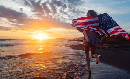 Young woman holding national American flag walking ocean beach. America Independence Day conceptの写真素材