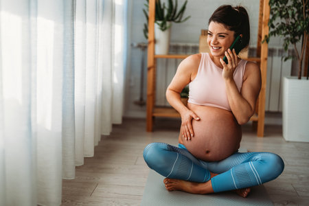 Pregnant sports woman taking a break from exercises and having phone call while relaxing at home