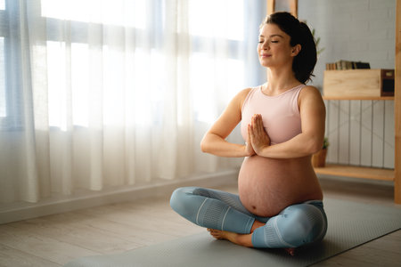 Fit pregnant woman doing breathing exercises while sitting cross-legged on an exercise mat at home