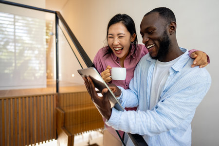 Multinational couple enjoying their morning routine, smiling and using laptop and tablet at homeの写真素材