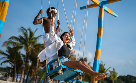 Happy interracial couple enjoying a fun beach day, swinging under palm trees on a tropical vacationの写真素材