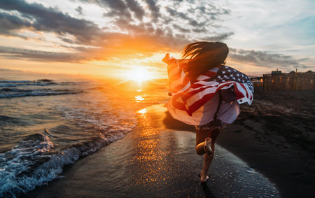 Young woman holding national American flag walking ocean beach. America Independence Day conceptの写真素材