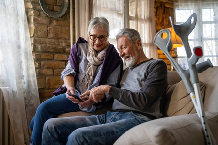 Senior wife embracing disabled husband at nursing home. Cheerful old couple using mobile phone.