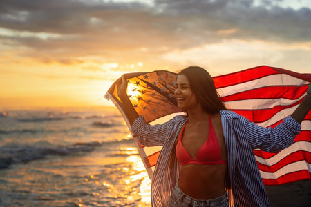 Beautiful patriotic young woman with the American flag held in her standing in the summer sunsetの写真素材