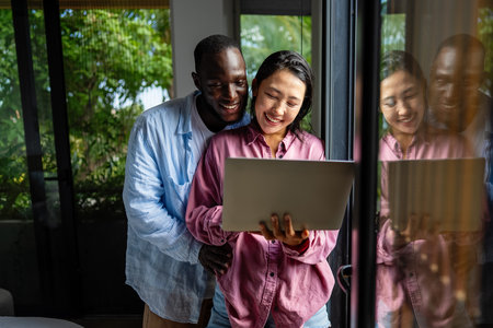 Happy black and Asian couple using laptop together, smiling wife and husband shopping onlineの写真素材