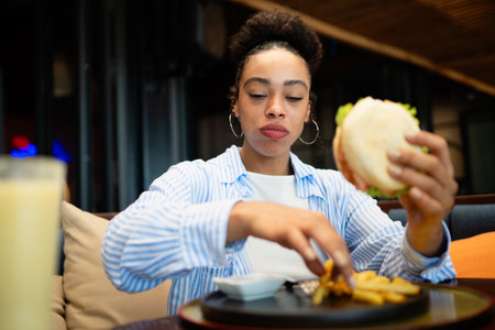 A moment of pure happiness. Smiling young woman savoring her burger and fries at a cozy restaurantの写真素材