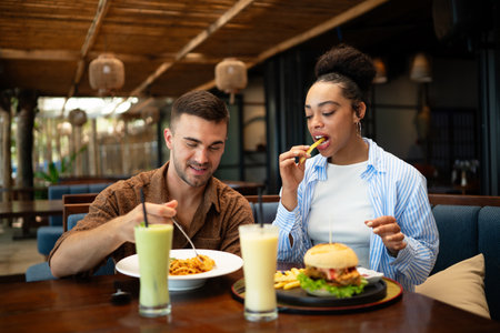 Happy diverse couple enjoying food at the restaurant, indulging in the guilty pleasure of fast food