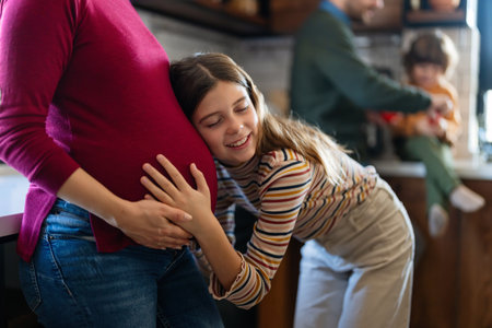 Happy daughter hugging belly her expecting mother at home. Excited girl listening to baby movementの写真素材