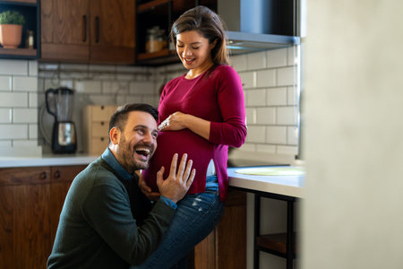 Beautiful pregnant woman and her handsome husband are smiling while spending time together at homeの写真素材