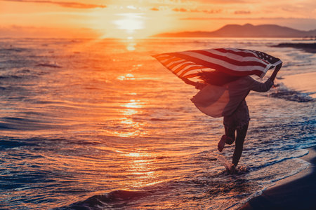 Beautiful patriotic young woman with the American flag held in her standing in the summer sunsetの写真素材
