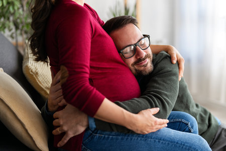 Beautiful pregnant woman and her handsome husband are smiling while spending time together at homeの写真素材