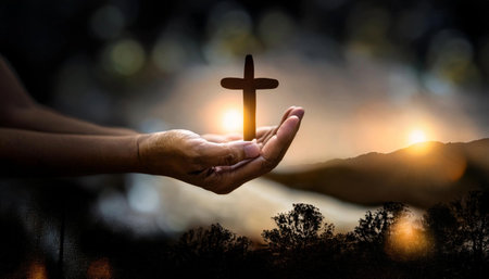 Hands holding a cross against sunrise over mountain range in the backgroundの素材