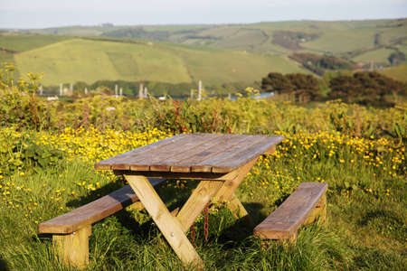 A picnic table surrounded by flowers with hills, a river and boats in the backgroundの写真素材