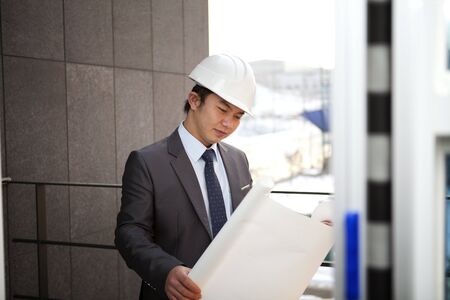 young architect with white helmet holding blueprint checking plan standing front office buildingの写真素材