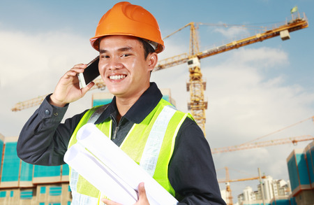 Portrait of an construction engineer holding blueprint convesation via phone with yellow crane and new building in the backgroundの写真素材