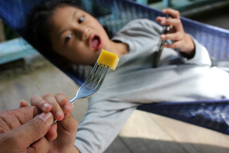 Dad feed a mango to his daughter who's only interested with the phone.の写真素材