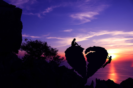 Women sit and man climbing on broken heart-shaped stone on a mountain with purple sky sunset background.Silhouette Valentine background concept.の写真素材