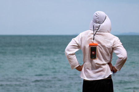 Tourists man wear a raincoat and hanging smartphones in the waterproof bag standing on the beach in Thailand.の写真素材