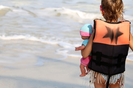 little girl stands holding a doll on the beach ocean waves background.の写真素材