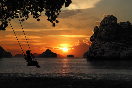 Silhouette of mother and son sitting on cradle or hammock on the beach with red sky sunset background.の写真素材