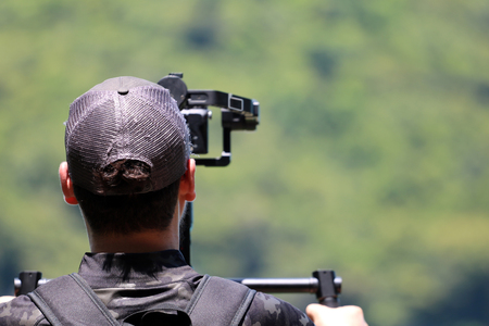 Photographer or videographer holding the video camera on a tripod on top of the mountain. Forest,ocean and blue sky  background.の写真素材