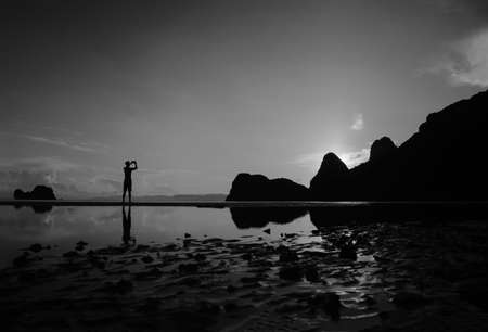 Man using a phone to take pictures of the morning sunrise on the beach, reflection on the water,...