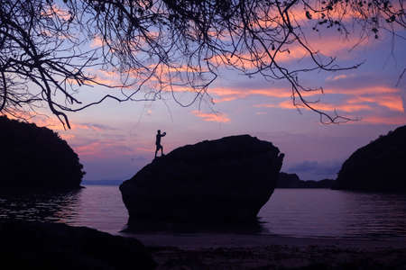 Man using a phone to take pictures of the morning sunrise on top of a rock on the beach at sunrise, morning sky blackground.の写真素材