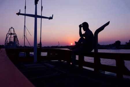 A man sits anxiously on a fishing boat parked by the canal at sunset in Thailand.の写真素材