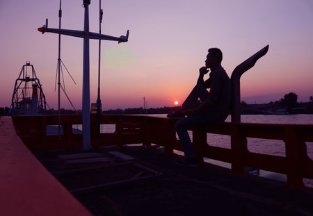 A man sits anxiously on a fishing boat parked by the canal at sunset in Thailand.の写真素材
