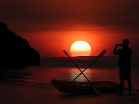 Silhouette of asian man stand and take sunset photo on beach near kayak in evening, Koh Samui, Thailand.の写真素材
