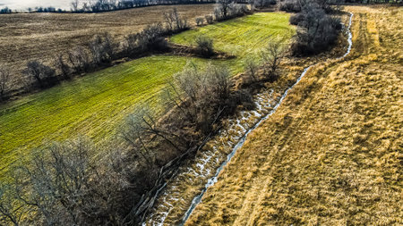 A frozen stream runs next to the agricultural fields with a grassy path as well.  Bare trees act as...
