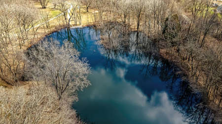 The trees are still bare just as winter has passed. The water is no longer frozen. Signs of fresh grass grow in the background. The sky is reflecting on the open waterの写真素材